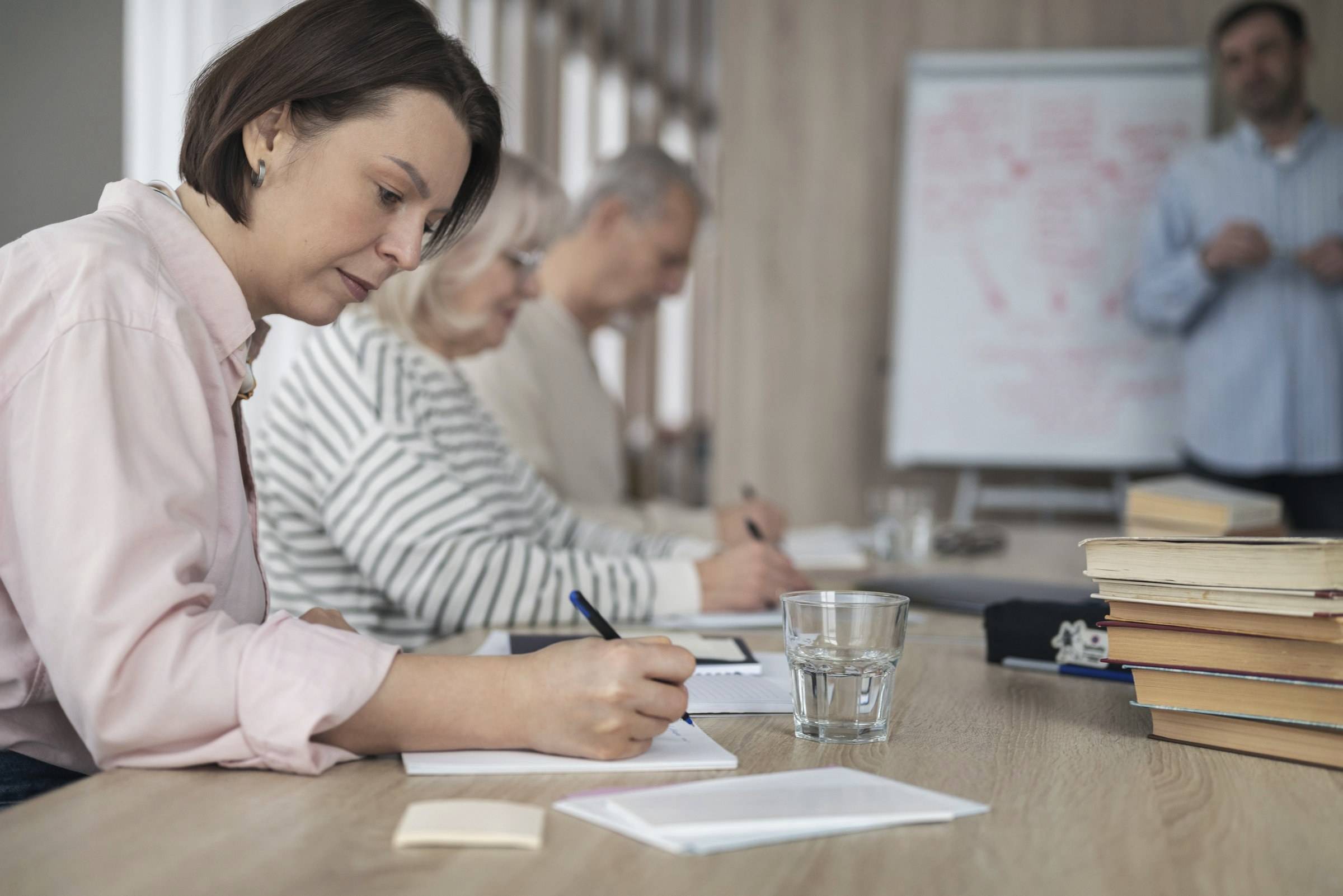 Erwachsene schreiben in einem Lehrgang an einem Tisch, vorne steht der Kursleiter am Flipchart